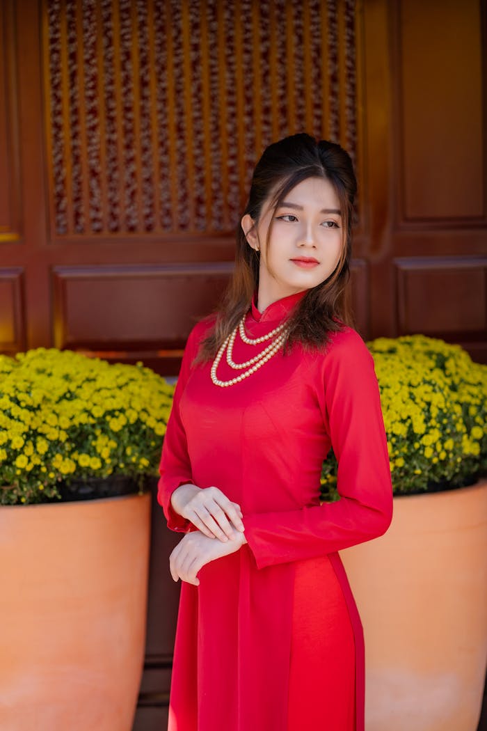 about-us Vietnamese woman in a red ao dai standing next to yellow flowers, symbolizing tradition and beauty.